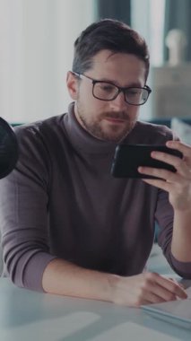 Şık gözlükler takan bir adam iletişim kurmak için aktif olarak cep telefonunu kullanırken rahat bir şekilde masada oturuyor.