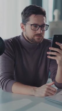 Gözlüklü bir adam cep telefonunu masada kullanıyor, konsantrasyonunu gösteriyor. Masasında bir mikrofon var, tartışmaları ve sunumları gösteriyor, teknoloji ve profesyonelliği harmanlıyor.