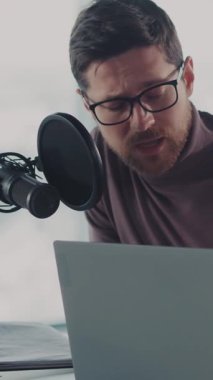 Gözlüklü bir adam dizüstü bilgisayar kullanırken mikrofona şarkı söylüyor, bu da müzik prodüksiyonunu gösteriyor. Daha sonra dijital projeler üzerinde dikkatle çalışır.