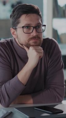 Gözlüklü ve sakallı bir adam, derin düşüncelere dalmış bir masada oturur, çenesini eline koyar, önemli kararları düşünür.