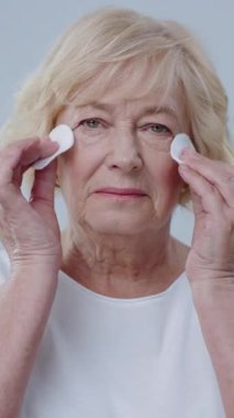 Yaşlı bir kadın nazikçe yüzüne çeşitli cilt bakım ürünleri uyguluyor ve kişisel bakımın önemini sergiliyor.