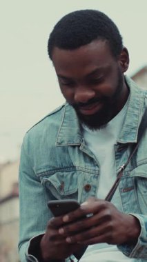 Şık bir kot ceket giyen genç bir adam, hareketli bir şehir ortamında akıllı telefonuyla etkileşime geçiyor.