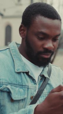 Bir adam şık bir kot ceket giyerken akıllı telefonunu kontrol eder, canlı bir şehir arka planına göre ayarlanır.