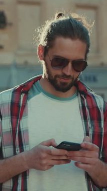 Güneş gözlüklü güler yüzlü genç adam telefon caddesinde yürüyor günlük kıyafet şehirleri geziyor güneşli güneş ışığına bakıyor genç mükemmel görünümlü yavaş çekim portresi yakın çekim