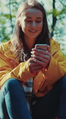 Sarı ceketli güzel bir kadın ormanda bir taşın üzerinde oturuyor, gülümsüyor ve akıllı telefonunu kullanıyor. İnternete göz atmak, mesajlaşmak, sosyal ağlar. Modern iletişim. Kadın portresi