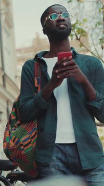 Şık ve şık bir adam akıllı telefonunu canlı ve hareketli bir şehir ortamında tutarken düşünceli bir şekilde durur.