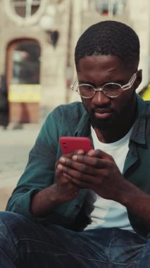 Dışarıda oturan genç bir adam, insanlar arka planda yürürken, akıllı telefonuna derinden odaklanıyor.