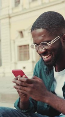 Gözlük takan genç bir adam açık hava ve modern teknolojinin tadını çıkarırken akıllı telefonuyla derinden meşgul.