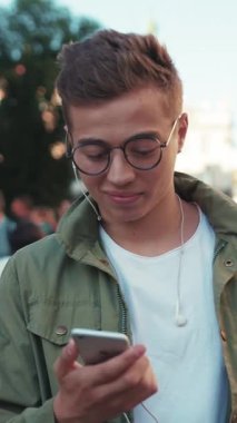 Gözlüklü genç adam telefon kullan. Kulaklıkla müzik dinle. Kameraya bak. Mutlu cep telefonu gülümse.