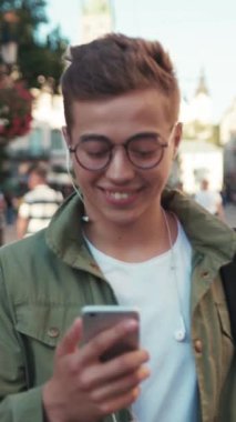 Gözlüklü hoş bir genç adam telefon kullanıyor kulaklıkla müzik dinliyor mutlu bir cep telefonu ışığı gezici cihaz dışarıda akıllı telefon teknolojisi yakışıklı hippi yavaş çekim portresini kapat