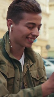 Yüzlü genç adam telefon kullanıyor. Kulaklıkla müzik dinliyor. Gülümsüyor, mutlu bir şekilde gülümsüyor.