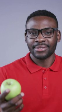 Süslü genç adam taze yeşil elma tutuyor ve gri arka planda gülümsüyor. Şık kıyafet, gözlük gibi inek. Diyet konsepti, fitness, spor. Erkek portresi, yakın görünüm