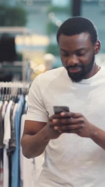 Genç bir adam, çağdaş bir alışveriş alanında rahatça dolaşırken akıllı telefonunu inceliyor.