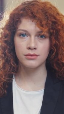 Kıvırcık saçlı dengeli bir genç kadın profesyonel bir stil ve düşünceli bir ifade sergiliyor..