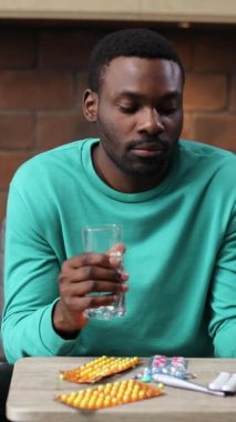 Sakallı hasta Afrikalı adamın portresi. Spor elbiseli çekici bir genç adam kanepeye oturmuş, ilaçlara bakıyor, elinde bir bardak su tutuyor. Tedavi.