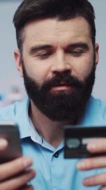Sakallı bir adam akıllı telefonu ve kredi kartıyla bağlanır, online alışveriş veya mali yönetimi gösterir, odaklanmış bir davranış gösterir.