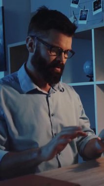 Gözlüklü sakallı bir adam masasında cep telefonuyla konuşuyor. İfadeleri, modern iletişimin görebilirliğini vurguluyor.