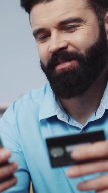 Mavi gömlekli sakallı bir adam elinde kredi kartı olan cep telefonunu kullanıyor. Onun odaklı ifadesi modern yaşam tarzı ve dijital finansmanı iletir