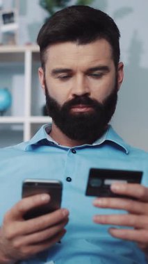 Bu videoda, sakallı bir adam koltuğundan online alışveriş yapıyor, telefonunu ve kredi kartını kullanarak, modern kolaylığı ön plana çıkarıyor.