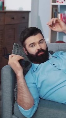 Bu videoda, sakallı bir adam bir kanepede dinleniyor, telefonunu ve uzaktan kumandasını kullanıyor. Modern hayatı yansıtan, teknolojiyle gelişigüzel etkileşimini yakalıyor.