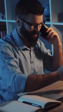 Mavi gömlekli sakallı bir adam masasında dizüstü bilgisayarı ve cep telefonuyla çalışıyor, odaklanmış ve ara sıra kollarını uzatıyor.