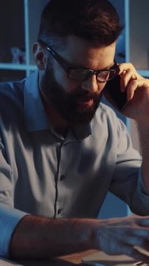 Gözlüklü bir profesyonel cep telefonuyla konuşuyor. Bilgisayarıyla birden fazla iş yapıyor. Teknoloji ve iletişimi karıştırıyor.