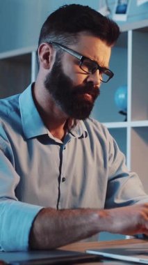 Modern bir çalışma alanında gözlüklü sakallı bir adam laptopuyla çalışıyor, verileri kontrol ediyor, önemli aramalar yapıyor, verimlilik sergiliyor.