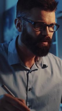 Bu videoda, gözlüklü sakallı bir adam teknolojik yeteneklerini tablet kullanarak gösteriyor, sonra da modern ve geleneksel eserleri harmanlayarak kağıda not alıyor.