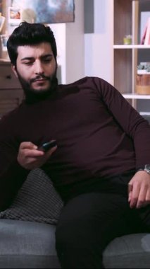 Bu rahat oturma odasında, sakallı bir adam rahat bir kanepeye uzanıyor, cep telefonu ve uzaktan kumandayla, peluş yastıklar ve battaniyeyle çevrili.