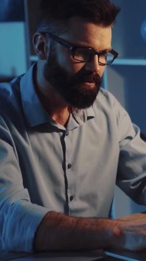 Sakallı ve gözlüklü neşeli bir adam evde günlük aktivitelere katılıyor, laptopuyla çalışıyor ve dişlerini fırçalıyor, neşeli kişiliğini sergiliyor.