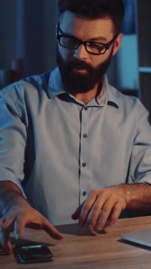 Gözlüklü ve mavi gömlekli sakallı bir adam bir masada dizüstü bilgisayar ve cep telefonuyla profesyonel görevleri ve kişisel görüşmeleri karıştırmakla meşgul.