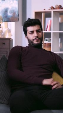 Sıcak bir oturma odasında, sakallı bir adam yastıklı ve battaniyeli bir kanepede dinlenir, cep telefonunu ve uzaktan kumandasını kullanır, keyfine bakar.