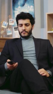 Takım elbiseli ve balıkçı yakalı şık bir adam oturma odasında bir kanepe ve şifoniyerle oturuyor. Kanepeden yere doğru kayıyor, telefonuyla meşgul oluyor.