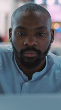 Mavi gömlekli düşünceli bir genç adam, parlak, modern ofis alanındaki dizüstü bilgisayarına odaklanmış durumda.