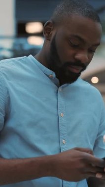 Şık mavi gömlekli düşünceli bir adam, akıllı telefonuyla modern bir ortamda iletişim kuruyor.