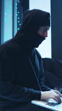 Kapüşonlu bir figür, dizüstü bilgisayara odaklanmış, teknolojiyi ve gizliliği temsil ediyor, büyüleyici, gizemli bir görsel yaratıyor.