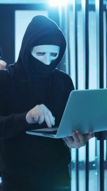 Gölgeli ve gizemli bir teknoloji ortamında kapüşonlu ve maskeli bir figür özenle dizüstü bilgisayarda çalışır.