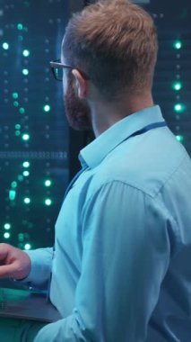 Yetenekli bir teknoloji uzmanı yoğun bir veri merkezinde özenle çalışıyor. Yüksek sunucu rafları arasında dizüstü bilgisayarına odaklanmış.