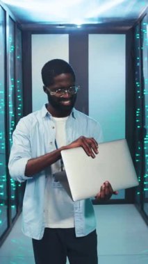 Teknoloji uzmanı gururla modern bir veri merkezinde duruyor. Parlak bir gülümsemeyle dizüstü bilgisayar gösteriyor.