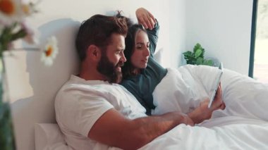 Pijamalı kaba sakallı bir adam karısını işaret ediyor. Tablet ekranında, yatakta uzanıp tartışırken ve gülerken. Sabah rutini, hafta sonu, rahatlama, tembellik..