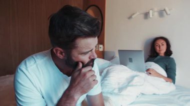 Sinirli sakallı adamın yataktan çıkışını yakından görüyor, yüzünü ve sakalını ovuyor, güzel karısı hala yatakta dizüstü bilgisayar kullanıyor. Çatışma, kötü ruh hali, sabah. Rahat yatak odası..