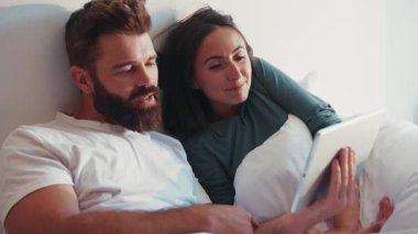 Genç çift pijamalarıyla yatakta yatarken tabletle internette sörf yapıyor, tartışıyor, yorum yapıyorlar. Tembel sabah, aktif kullanıcı, online alışveriş. Moda ailesi.