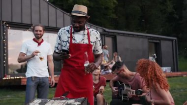 Yazlık şapkalı ve kırmızı önlüklü gülümseyen adam bira içer, akıllı telefonunu ızgarada yemekleri hazırlarken kullanır, arkadaşları şarkı söylerken, dans ederken, gitar melodileri dinlerken. Eğleniyor, yaz tatili.