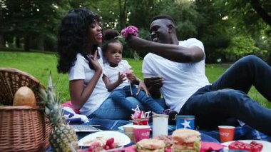 Gülümseyen Afro-Amerikan çift ve kızı şehir parkında genç bir adam çiçek sunuyor kadın ve çocuk piknik yapıyor dışarıda yavaş çekimde eğleniyorlar yaz ilişkisi kuruyorlar çocuk doğası birlikte rahatlayın
