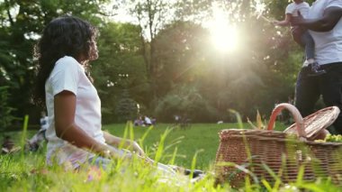 Günbatımı Afro-Amerikan çifti ve kızı şehir parkında oturmuş piknik yapıyorlar. Kadın ve çocuk piknik yapıyorlar. Dışarıda yavaş çekimde eğleniyorlar.