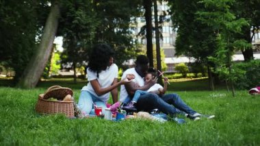 Ağır çekimde Afro-Amerikan ailesi parkta mutlu bir gülümseme yiyor. Genç kadın çocuk piknik yapıyor. Yavaş çekimde eğleniyor.