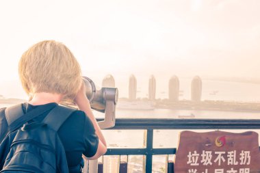 Sanya, Hainan adası, Çin - 16 Mayıs 2019: Bir kız dürbünle Sanya Phoenix Adası'nın panoramasını izliyor. Gün batımı, bulanıklık, pus.