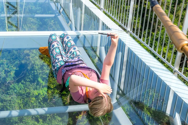 Hainan, Cina - 17 maggio 2019: Una giovane ragazza, sdraiata su un pavimento trasparente del ponte di osservazione sotto forma di ponte di vetro, si fa un selfie. Dietro il vetro c'è la giungla del parco forestale di Yanoda — Foto Stock