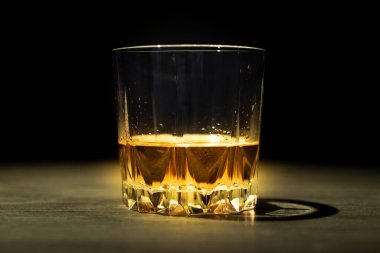 Bir ışık demeti tarafından aydınlatılmış bir bardak viski karanlık bir masada ve siyah bir arkaplanda duruyor.