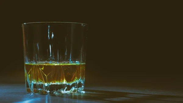 A glass of whiskey lit by a ray of light stands on a table, black background, copy space